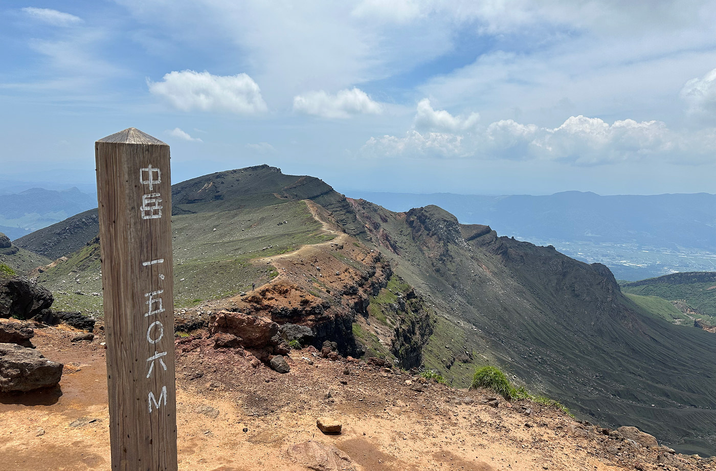 I förgrunden en påle med japanska tecken. I fjärran en lummig bergsås med gulbrun promenadled.
