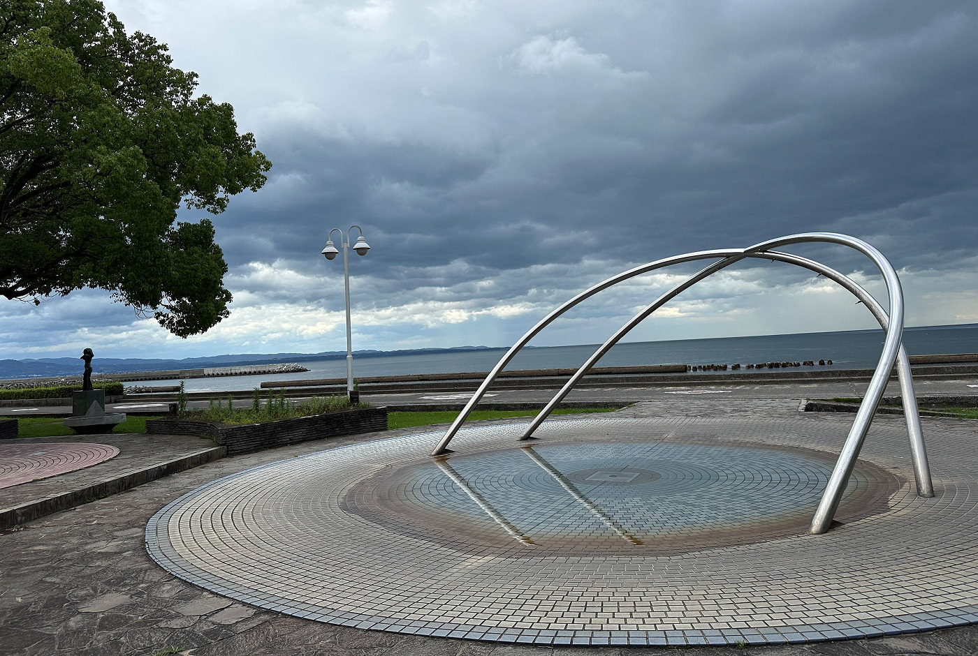 Skulptur med två böjda järnrör över en rund pool vid vatten nere vid en hamnpromenad.
