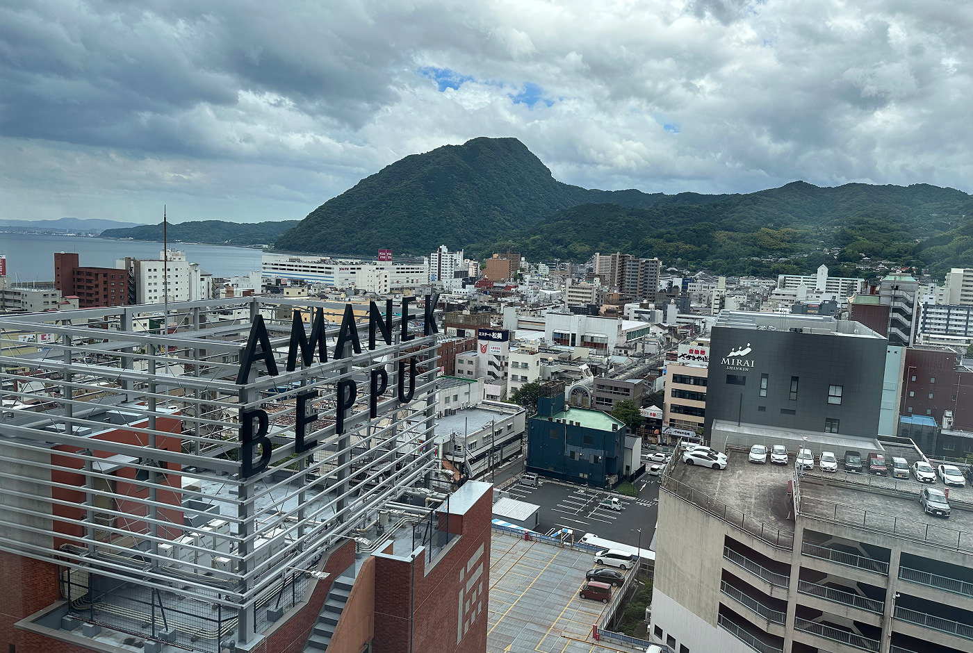 Utsikt över en stor hamnstad med ett lummigt berg i fjärran. En skylt på närmsta huset lyder "Amanek Beppu".