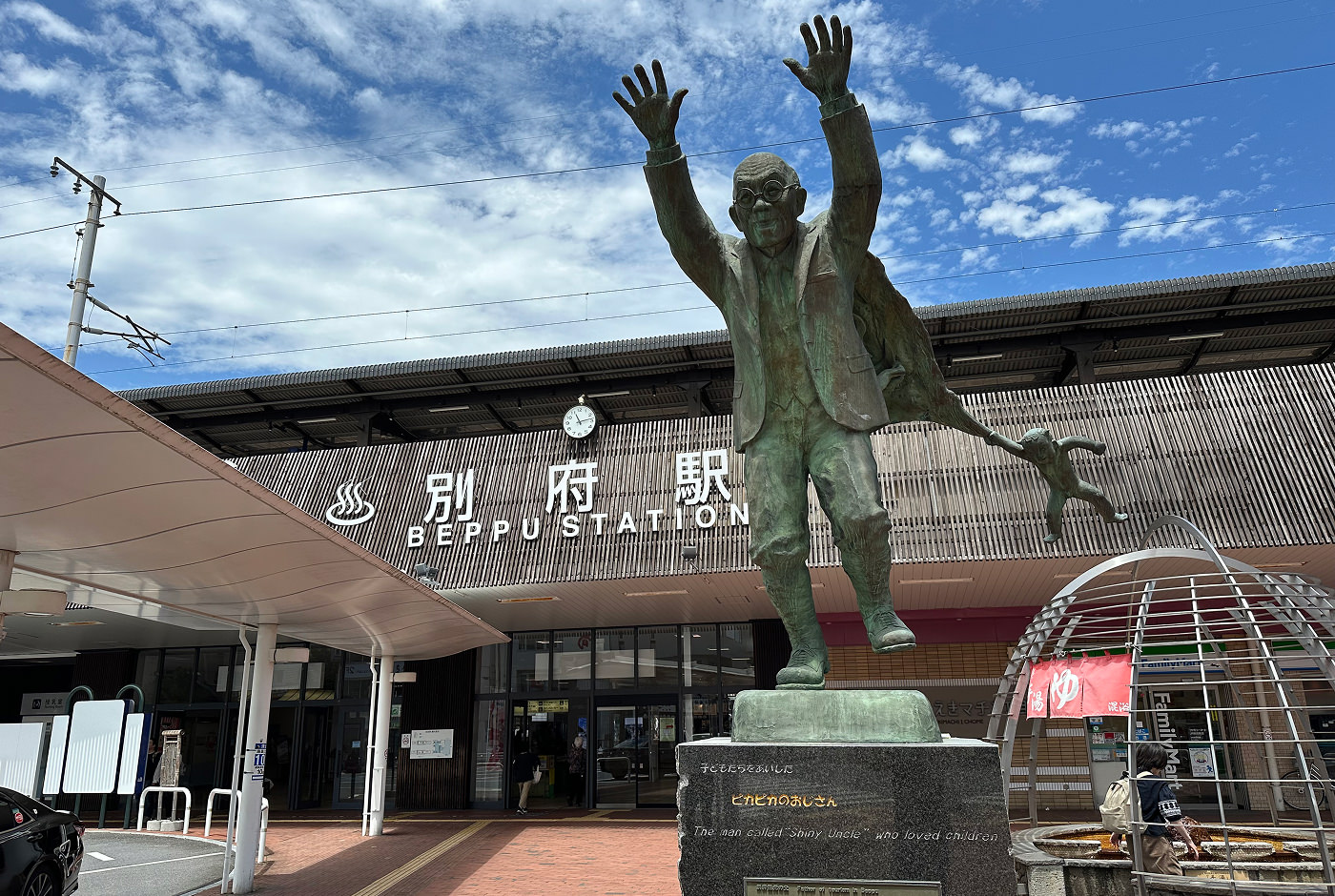 Staty på en gammal man med händerna i luften. Ett barn hänger i hans kappa. Står framför en tågstation.