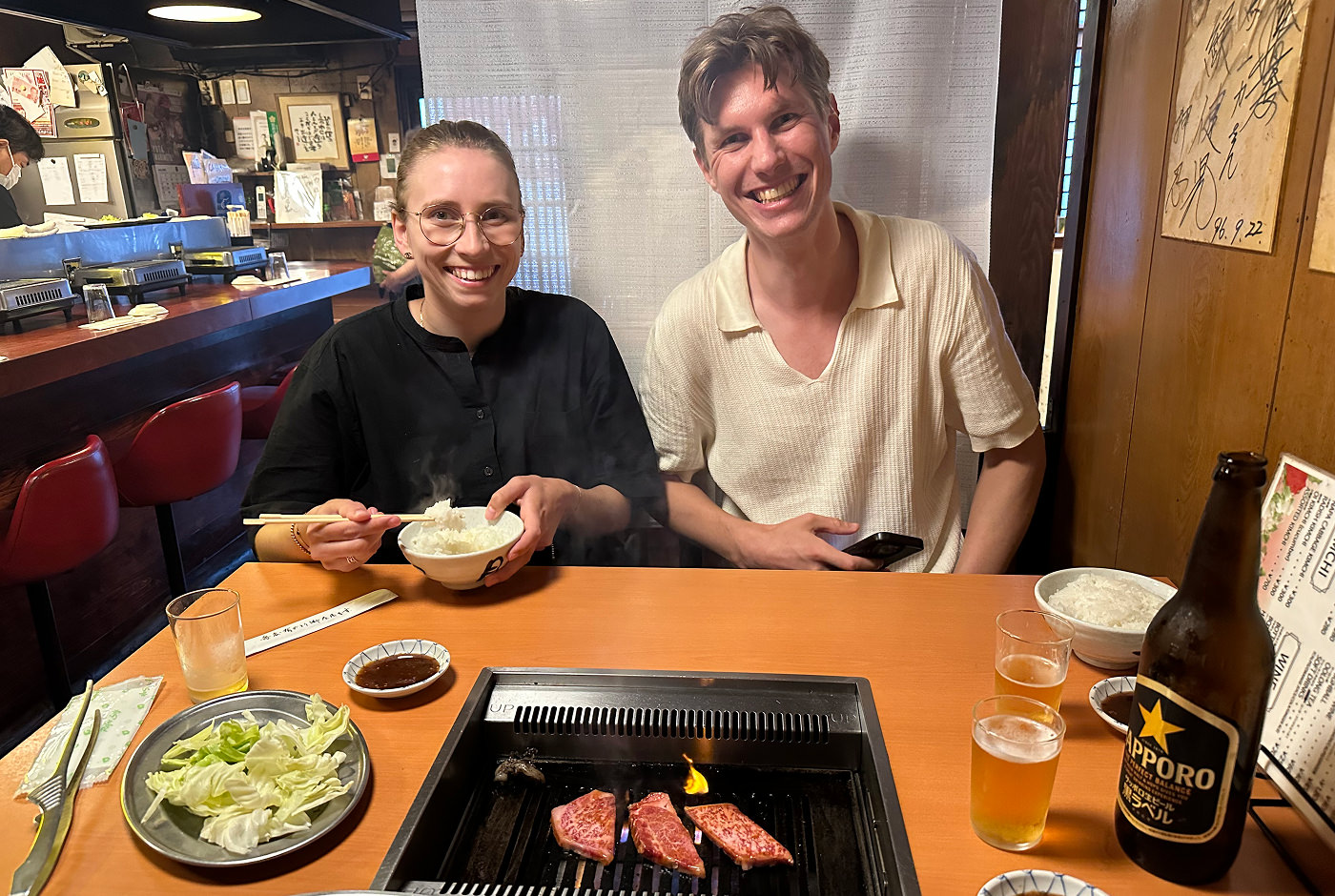 En ung kvinna och en ung man ler brett in i kameran samtidigt som de sitter och äter yakiniku.