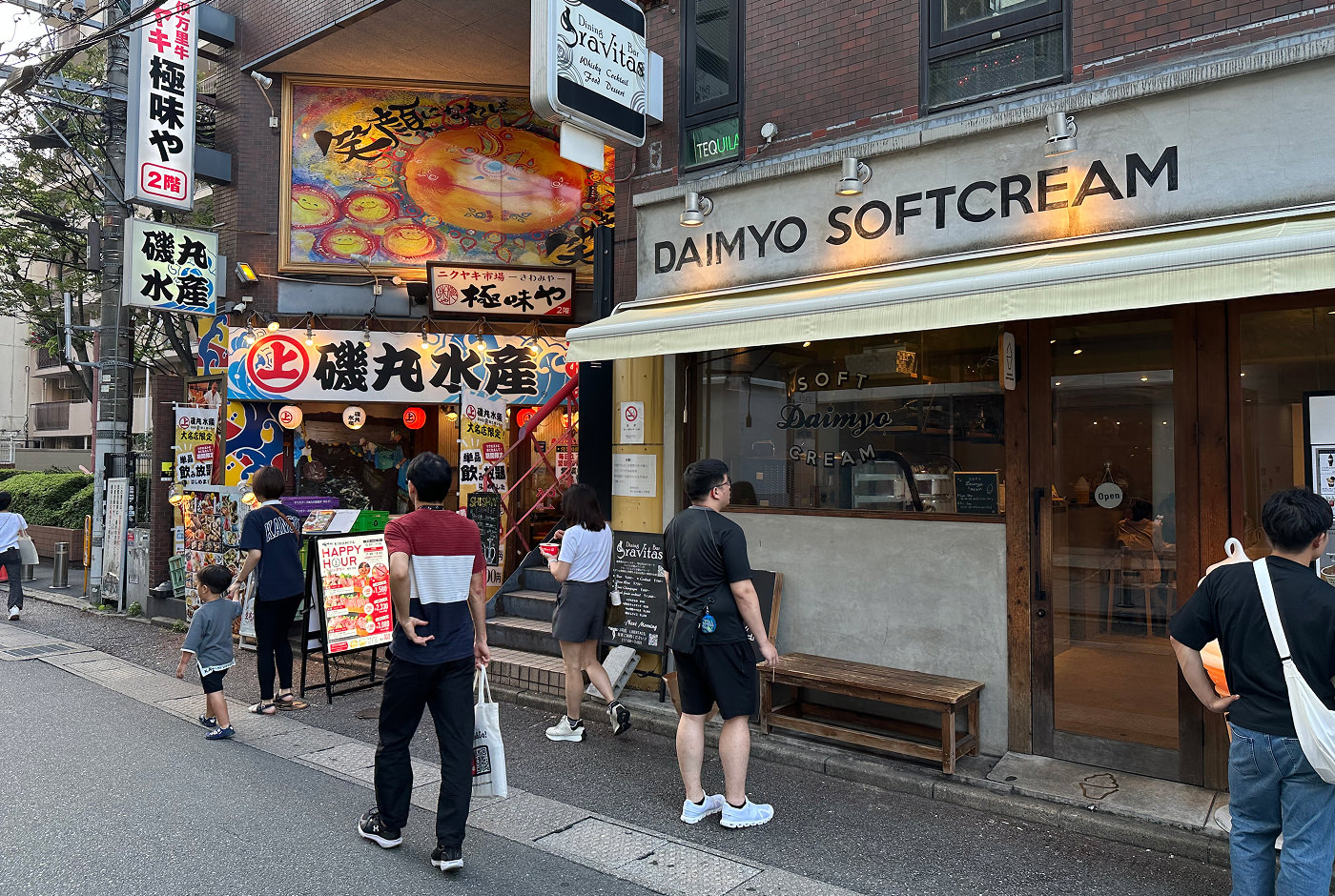 Sju personer går och stor vid en gata med två butiker. En fasad är färggrann med japansk skrift och en annan ljusgrå med texten "Daimyo Softcream".