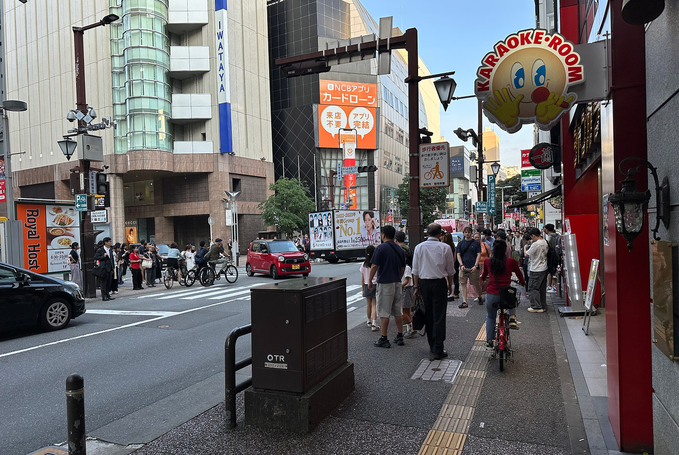 Korsning i en upptagen del av en stadskärna. Trottoarerna är fulla med folk och bilar kör på gatan. En skylt i förgrunden består av ett leende ansikte och texten "Karaoke Room".
