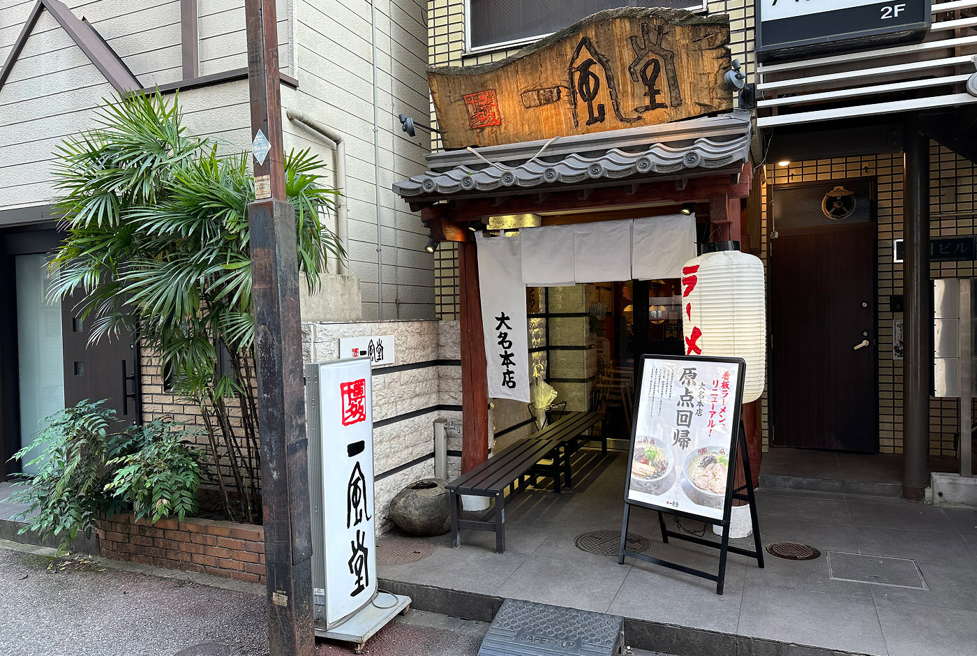 Ingång till en liten restaurang. Ovanför dörren finns en upplyst träskylt med japansk skrift. Vid dörren finns vita tygstycken, en bit lampa och en vit affisch med japansk skrift.