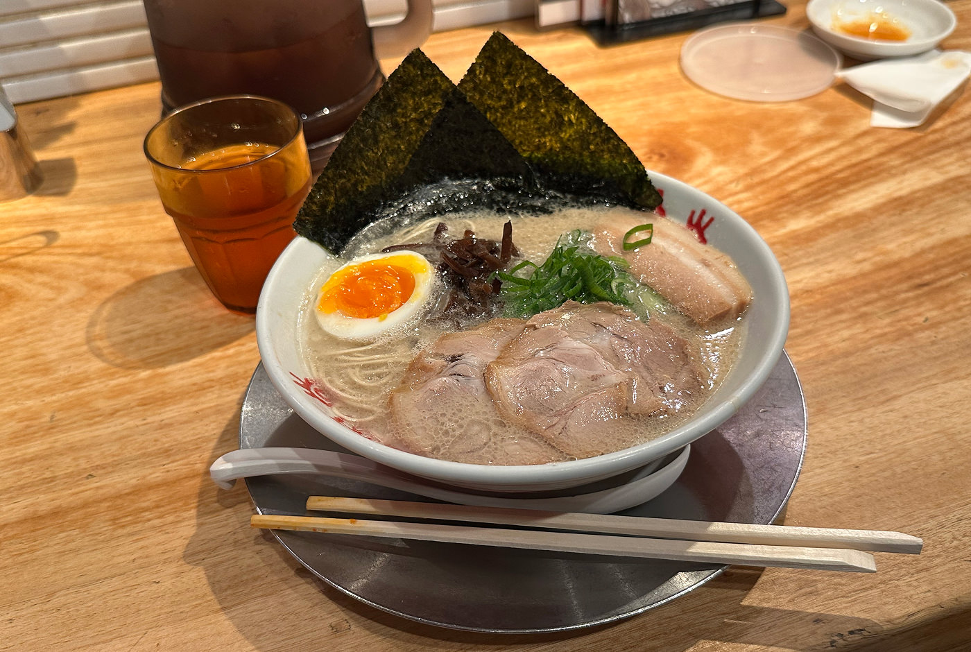 En vit skål med ljus ramen med ett löskokt ägg, sjögräs och fläskkött. Skålen står på en bardisk tillsammans med ett glas vatten.