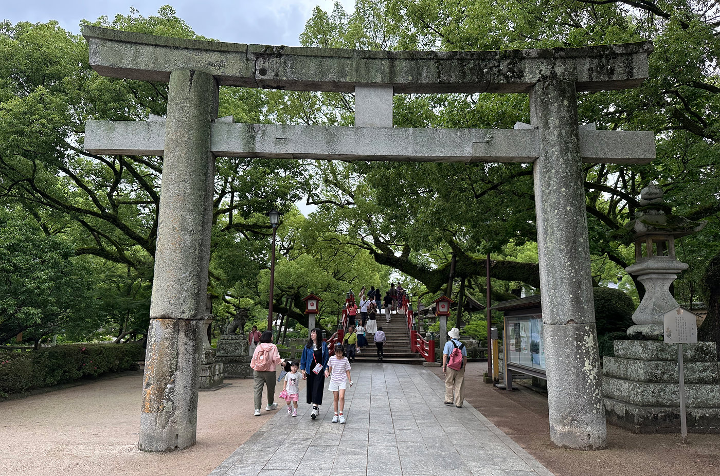Stor grå torii-port i sten omgiven av lummiga träd. Bakom den promenerar folk till en bra med röda räcken.