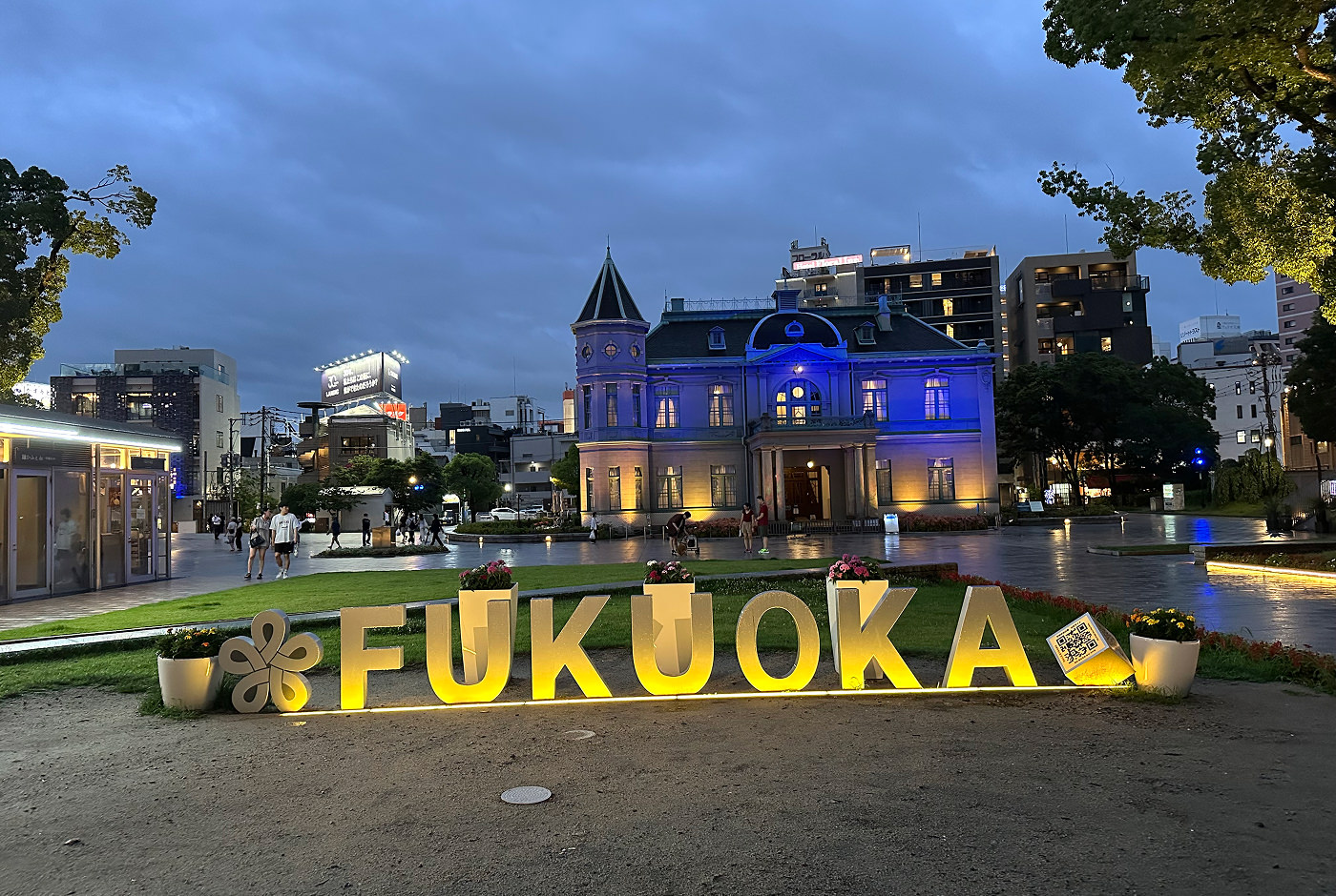 En upplyst gul skylt på marken som lyder "Fukuoka". Bakom den finns ett öppet torg och ett gammalt medborgarhus.