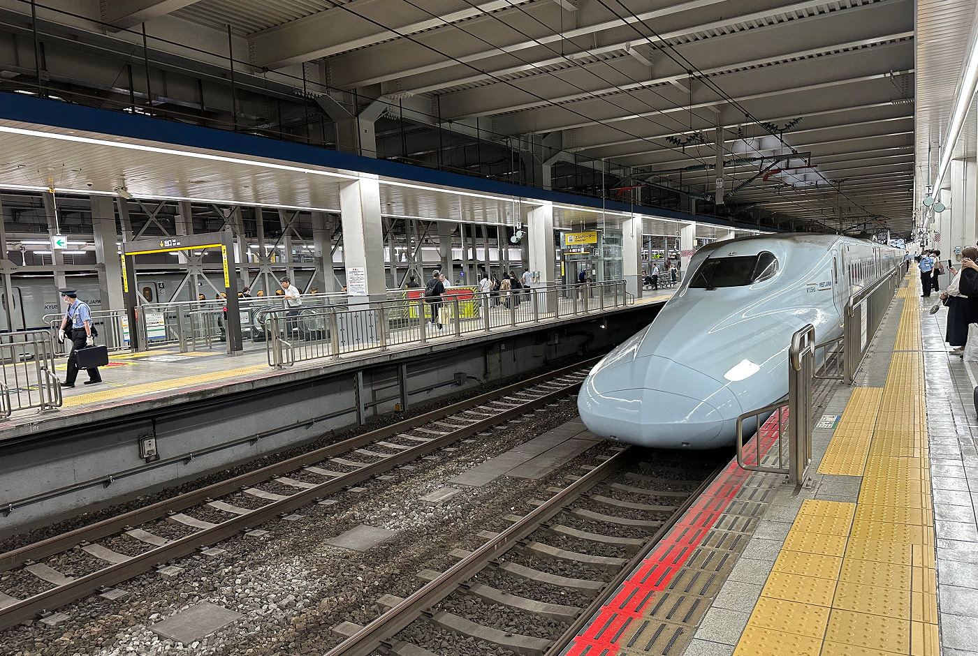 Lång tågperrong under tak. Ett långt Shinkansen-tåg håller på att stanna.
