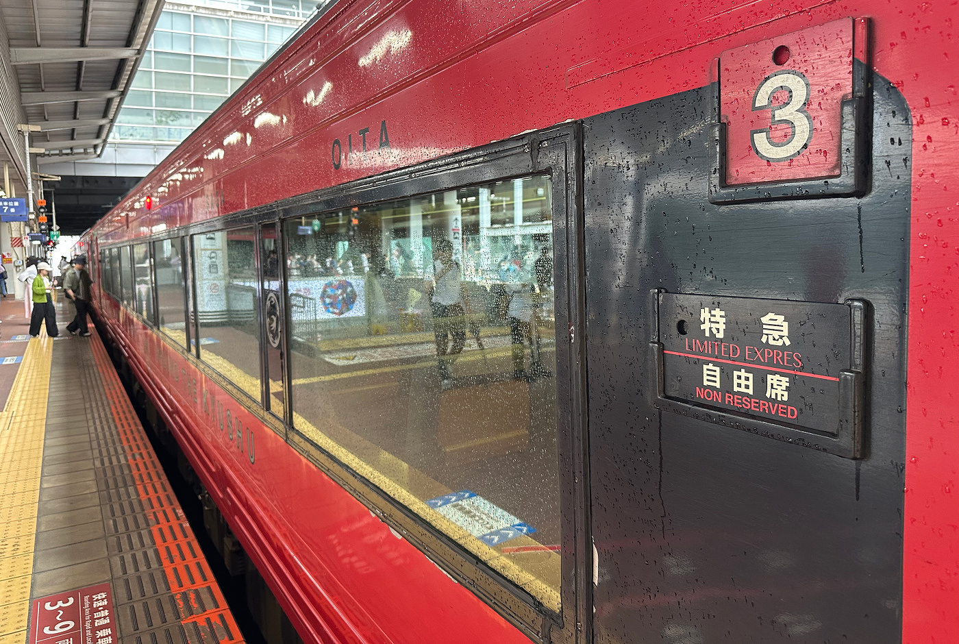 Röd tågvagn vid en perrong. Det har regnat på vagnen. Den har siffran "3" och texten "Limited Express Non Reserved".