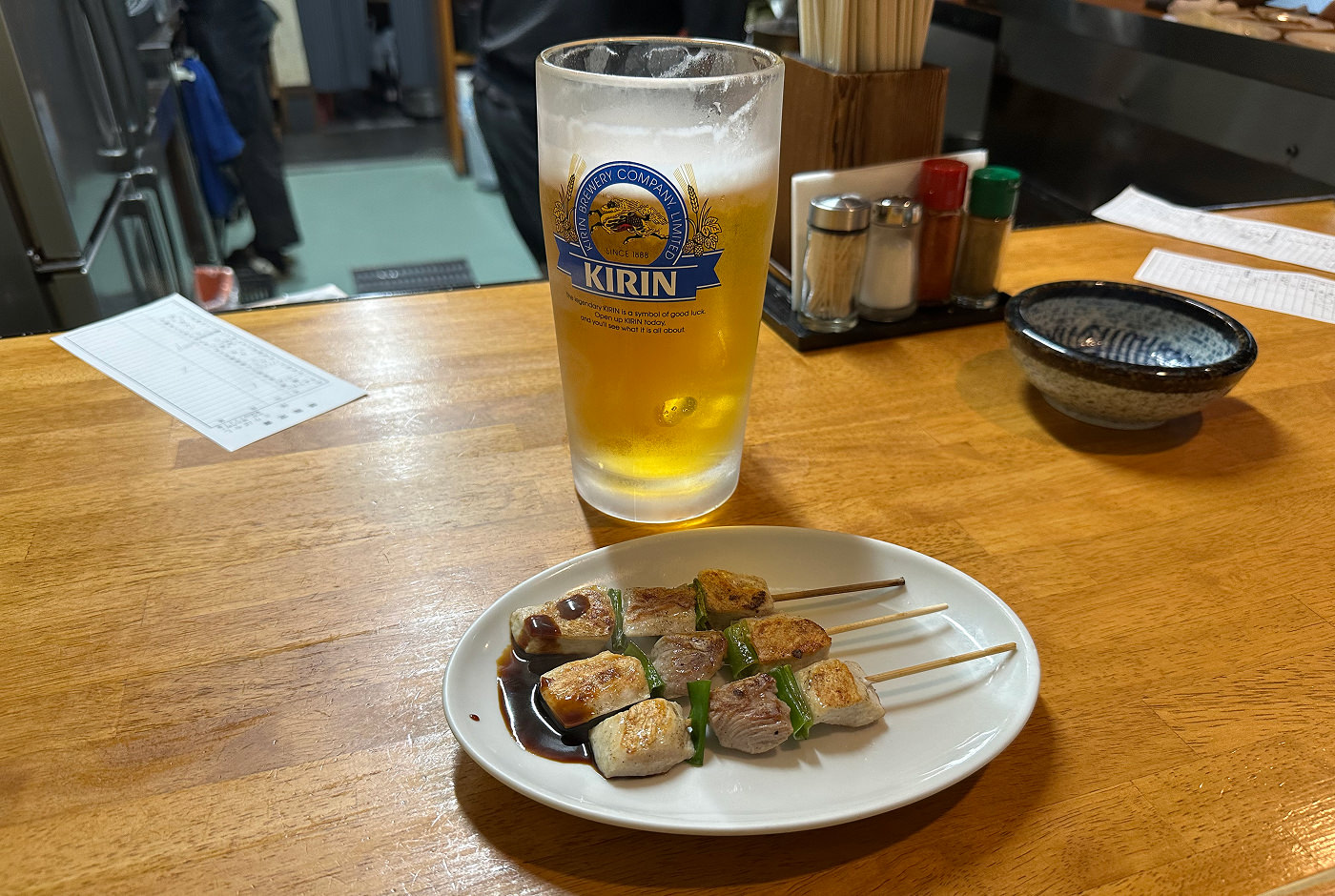 En bardisk med ett stort stop öl och en vit tallrik. På ölglaset står det "Kirin" och på tallriken ligger tre kycklingspett.