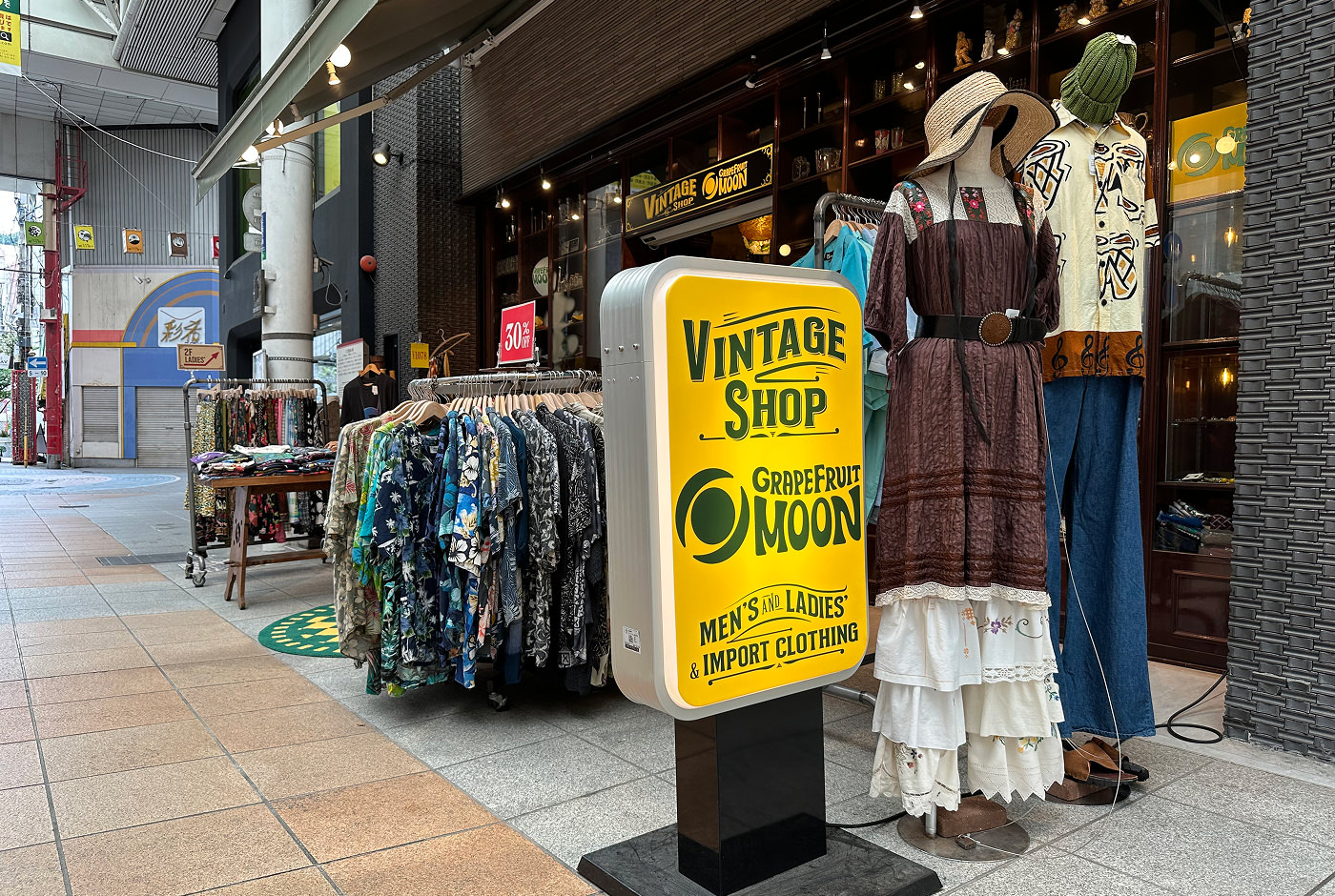 En gul skylt med grön text står på marken och lyder "Vintage Shop Grapefruit Moon" framför två skyltdockar med 70-talskläder bakom finns mer kläder och fasaden till butiken.