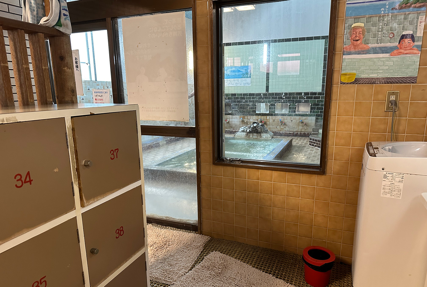 Omklädningsrum med skåp och tvättmaskin i ett litet badhus. Via ett fönster kan man se badet som har en rektangulär pool i mitten.
