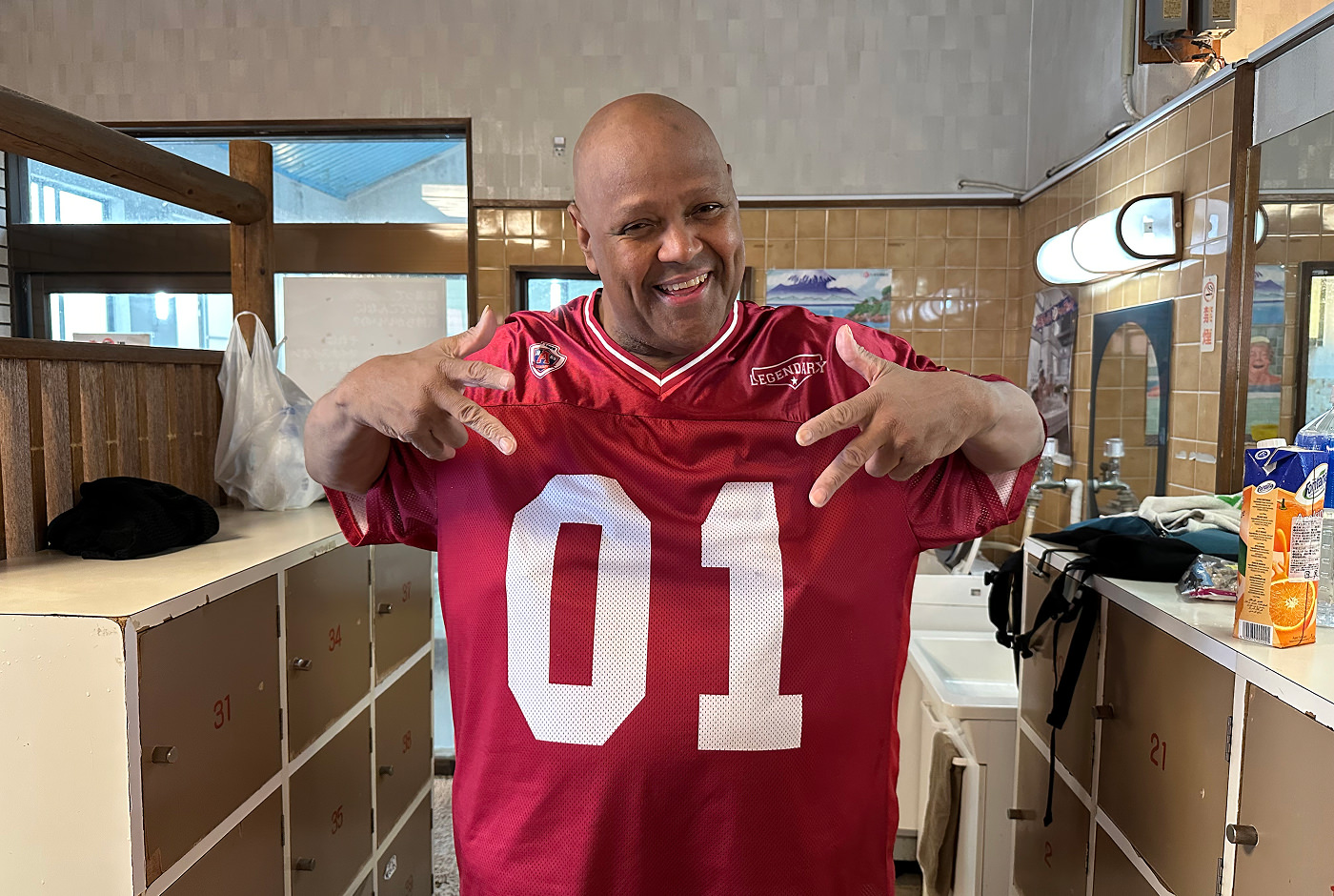 Glad afroamerikansk man med en vinröd sporttröja med texten "01" ler och poserar framför kameran i ett omklädningsrum.