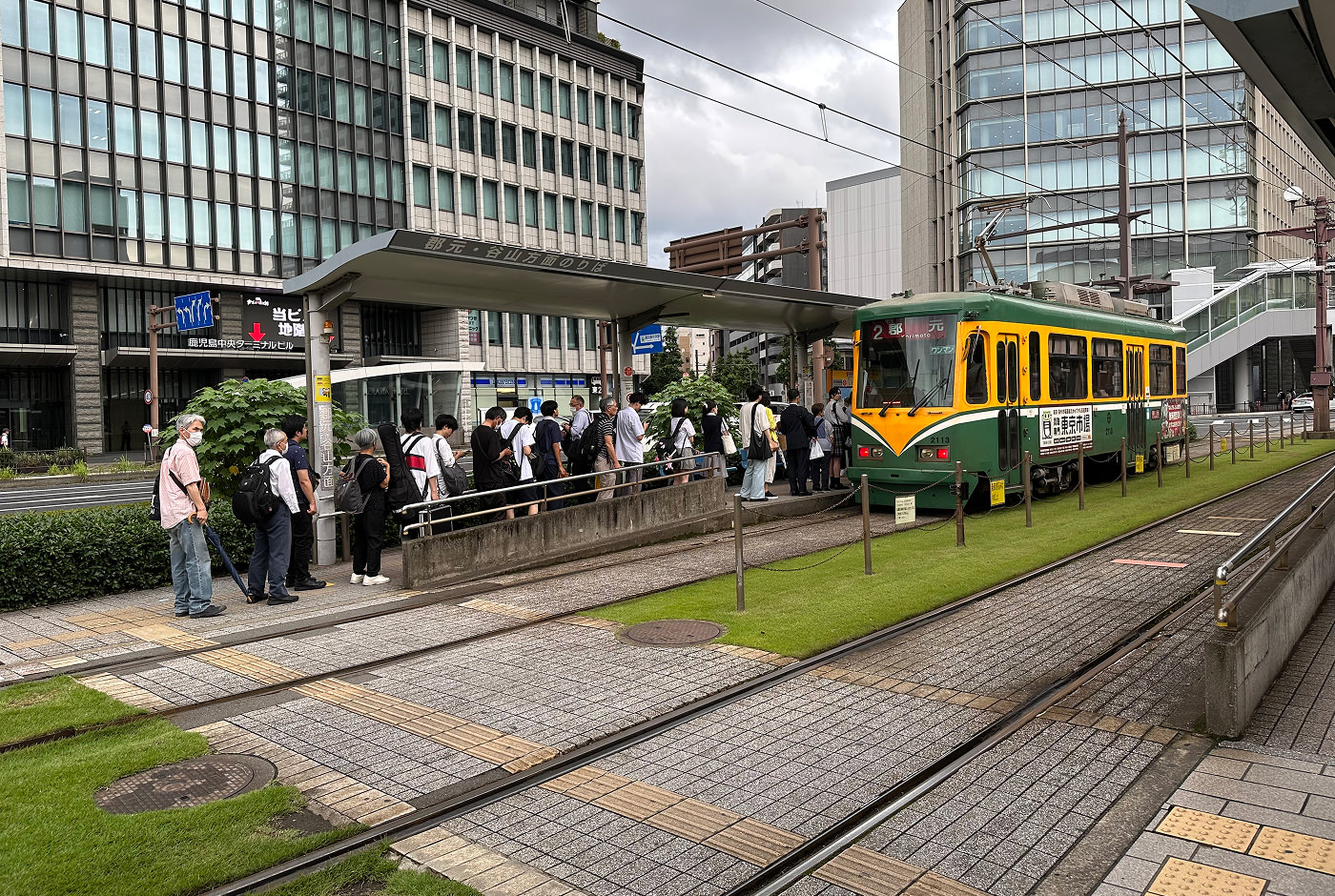Cirka 20 personer köar till en grön och gul spårvagn. Marken runtom består av välansat gräs och stenplattor. I bakgrunden finns höghus.