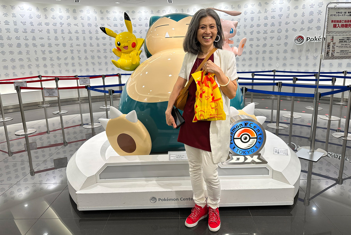 Keiko håller i en gul påse och står framför en stor staty över tre pokomons. En skylt lyder "Pokemon Center Tokyo".