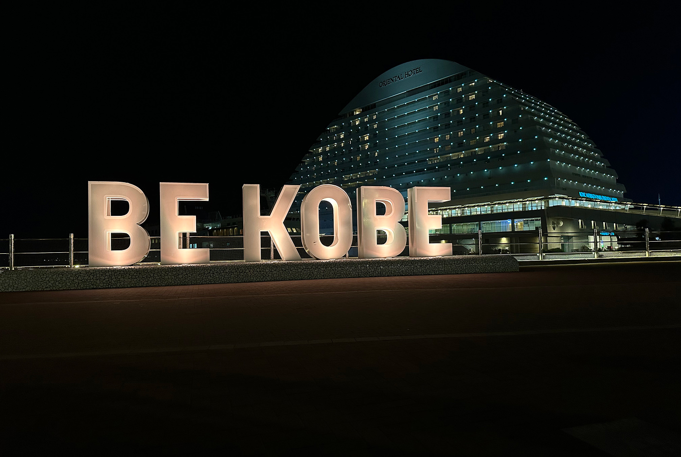 Fyra stora bokstäver som bildar texten ”Be Kobe”. Bokstäverna är upplysta. I bakgrunden står ett stort och upplyst ovalt hotell.