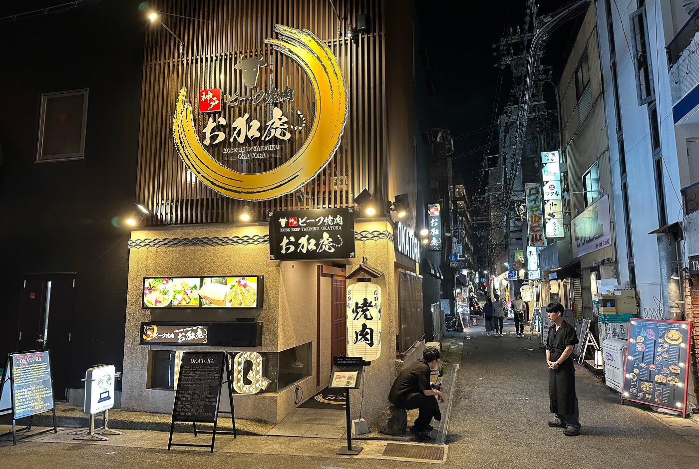 Två svartklädda servitörer står vid en upplyst gatukorsning utanför en restaurang med en stor skylt som består av ett gultrunt penseldrag och texten "kobe Beef Yakiniku Okatora"inuti. Det är mörkt ute.