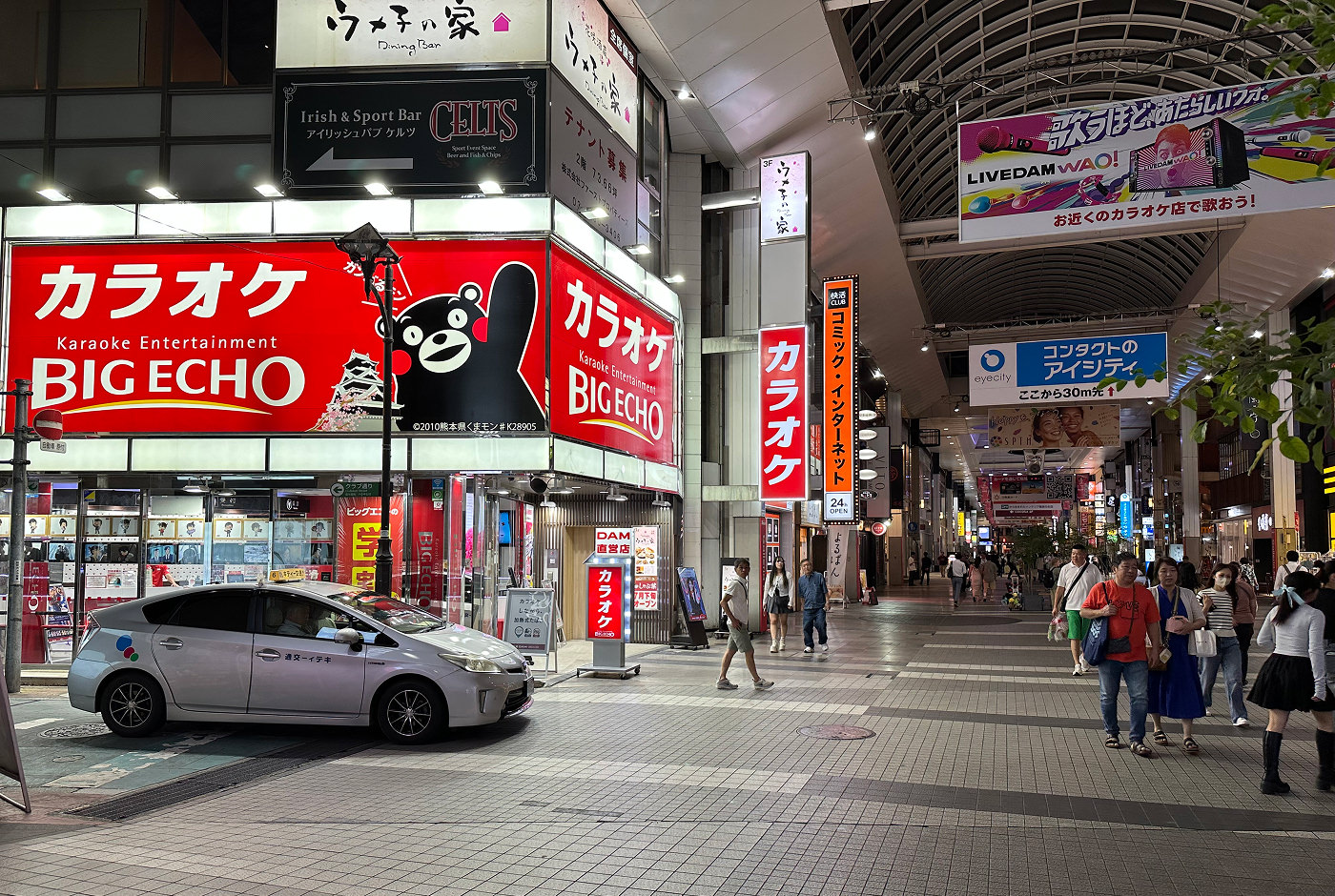 Den svarta glada björnen Kumamon syns på en stor röd reklamskylt med texten "Big Echo" på sidan av en låg shoppingarkad.