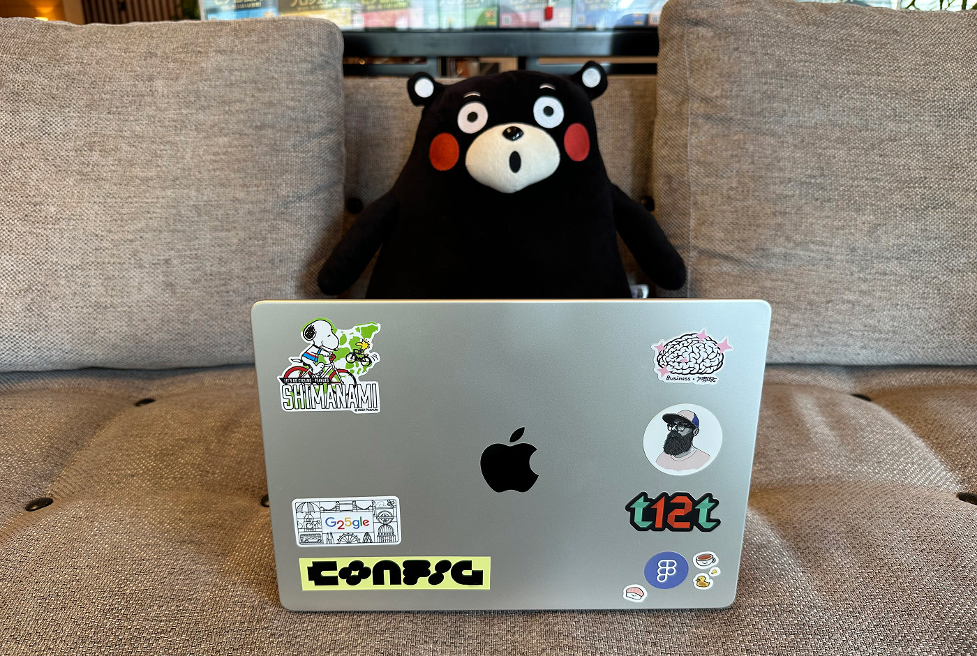 En barbar dator med klistermärken och uppvikt skärm står på en soffa framför det svarta och förvånade björnmonstret Kumamon.