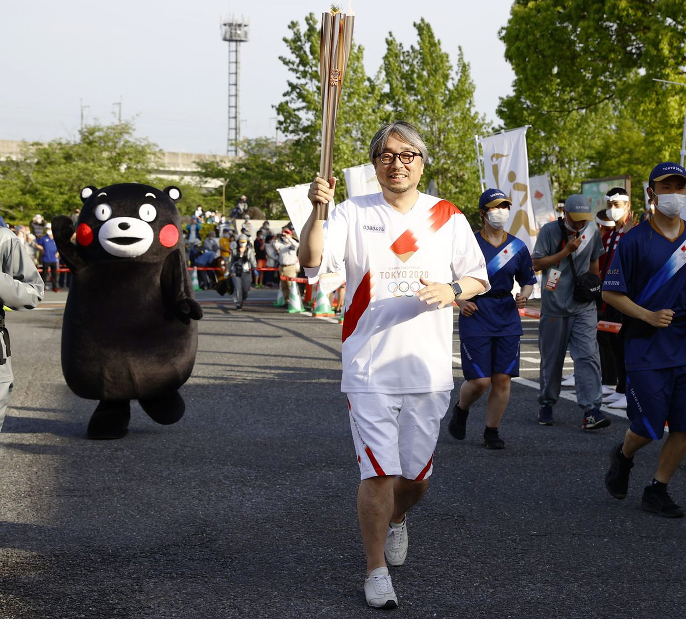 En japansk man med glasögon och ljusa sportkläder springer med den olympiska facklan. Bakom honom springer björnmonstret Kumamon.