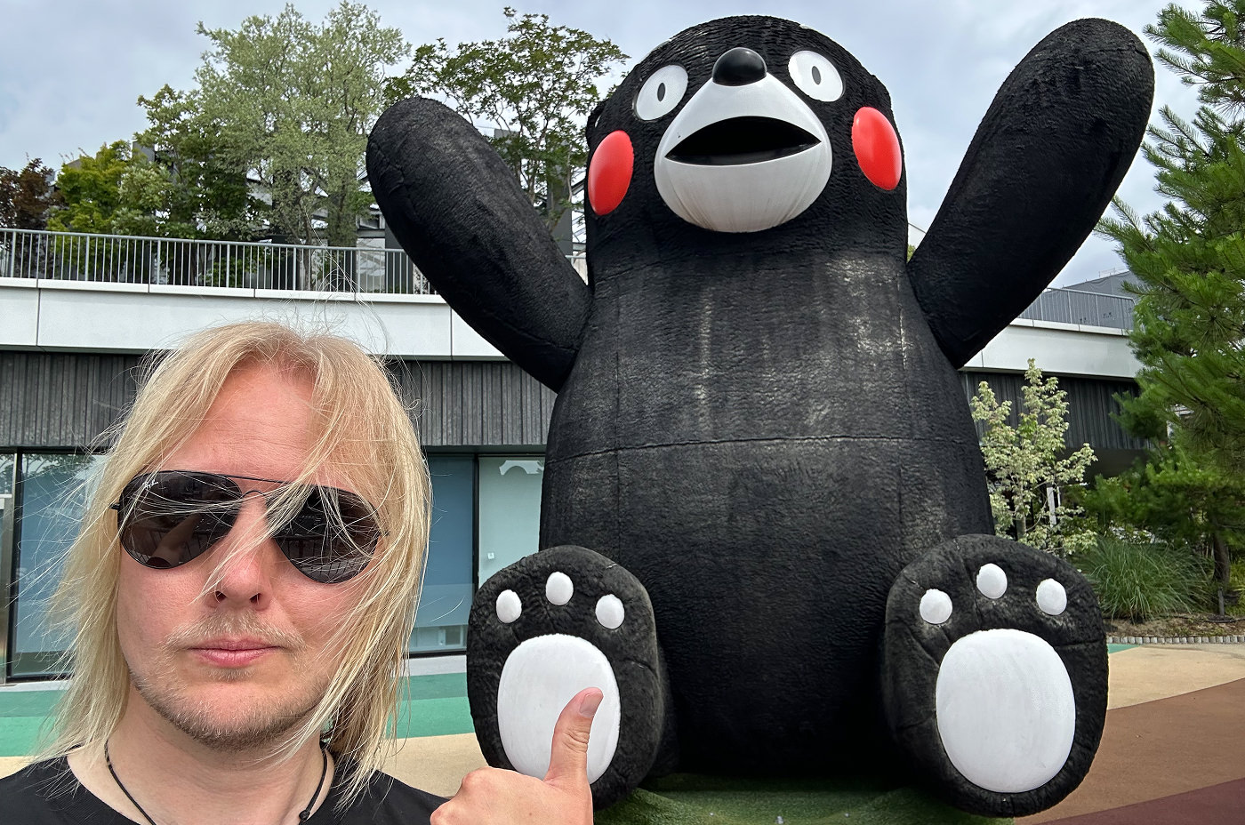 Jag har solgasögon på mig och tittar in i kameran. Med tummen pekar jag på en stor staty av den svarta glada björnen Kumamon.