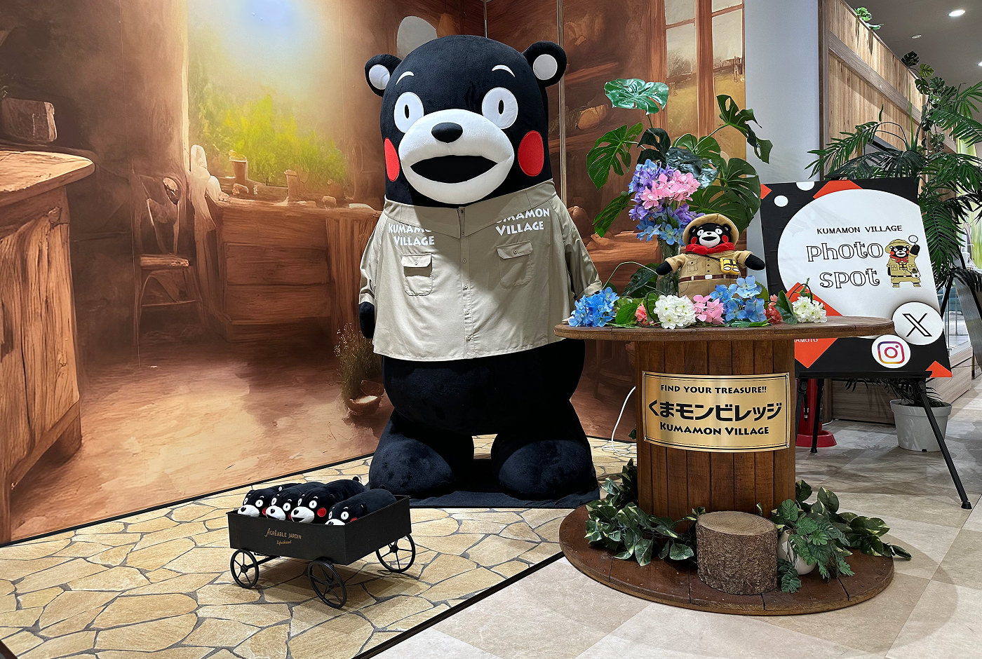 Stor staty av det glada björnmonstret Kumamon framför en plansch av ett mysigt trärum. Till höger står ett bord med en Kumamon-kramdjur, på golvet stor en liten kärra med tre kramdjur till.