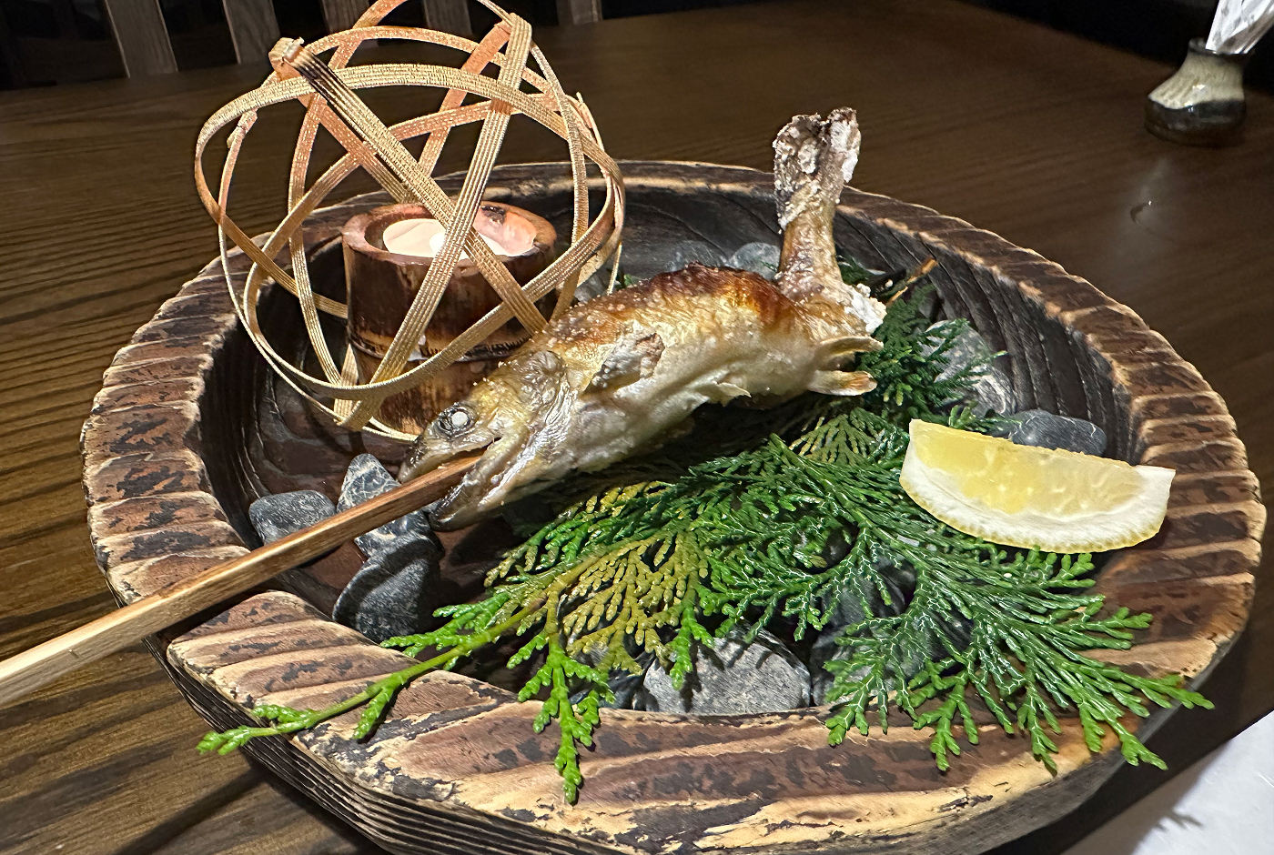 En grillad fisk med ett spett genom munnen på en trätallrik med gråa stenar, ett ljus och en citronklyfta.