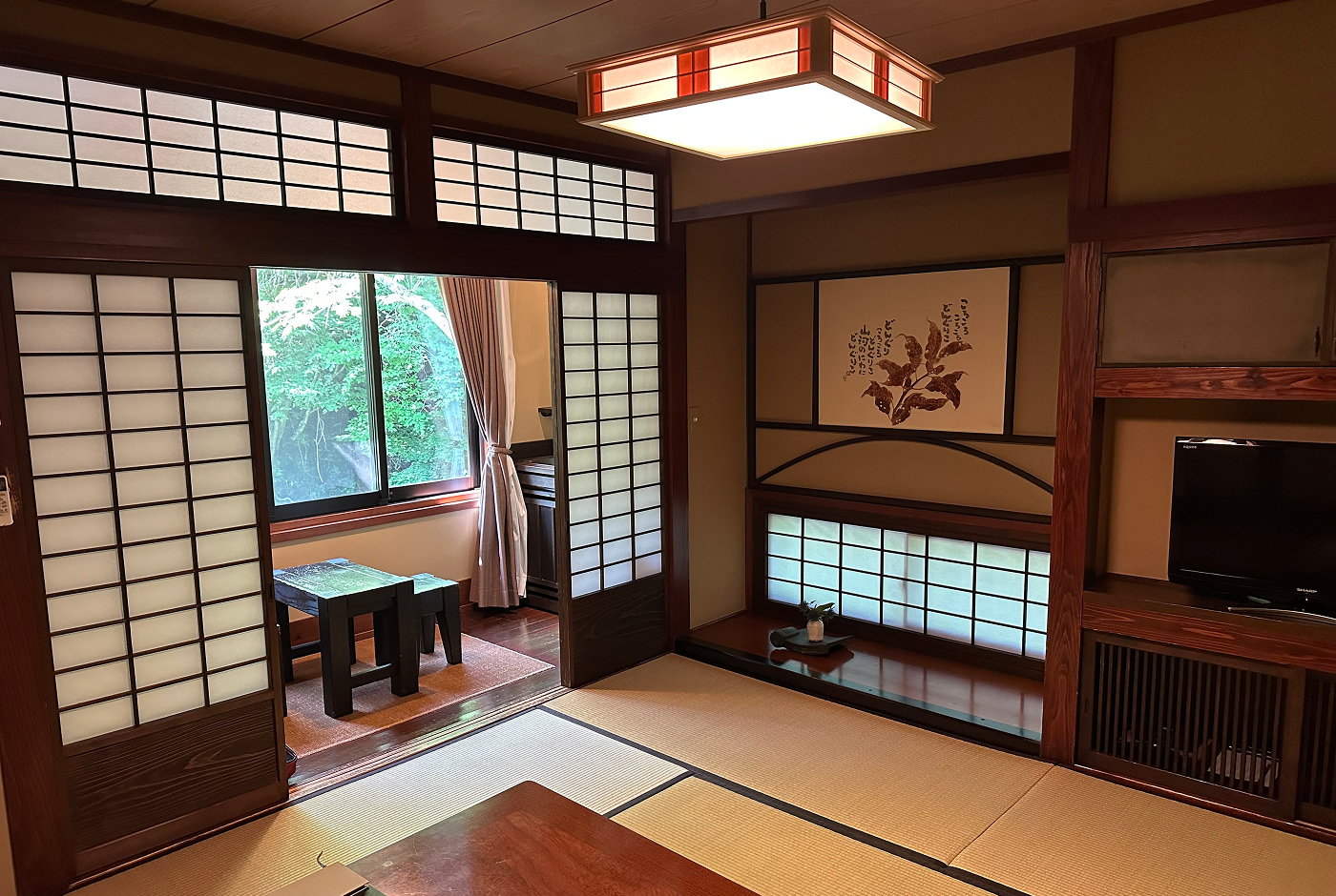 Rum i en traditionell ryokan med ljusa tatami-mattor på golvet. På väggen hänger en tavla av en växt. Bakom två skjutdörrar ser man ut i skogen via ett fönster.