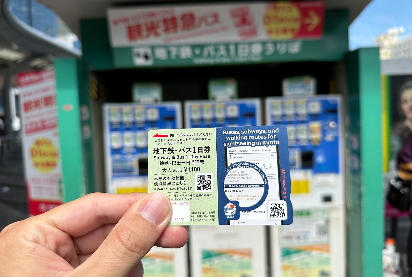 Hand håller i en blågrön pappersbit med mycket text på japanska och engelska. En del av texten lyder "Subway & Bus 1-Day Pass". I bakgrunden står tre biljettmaskiner.