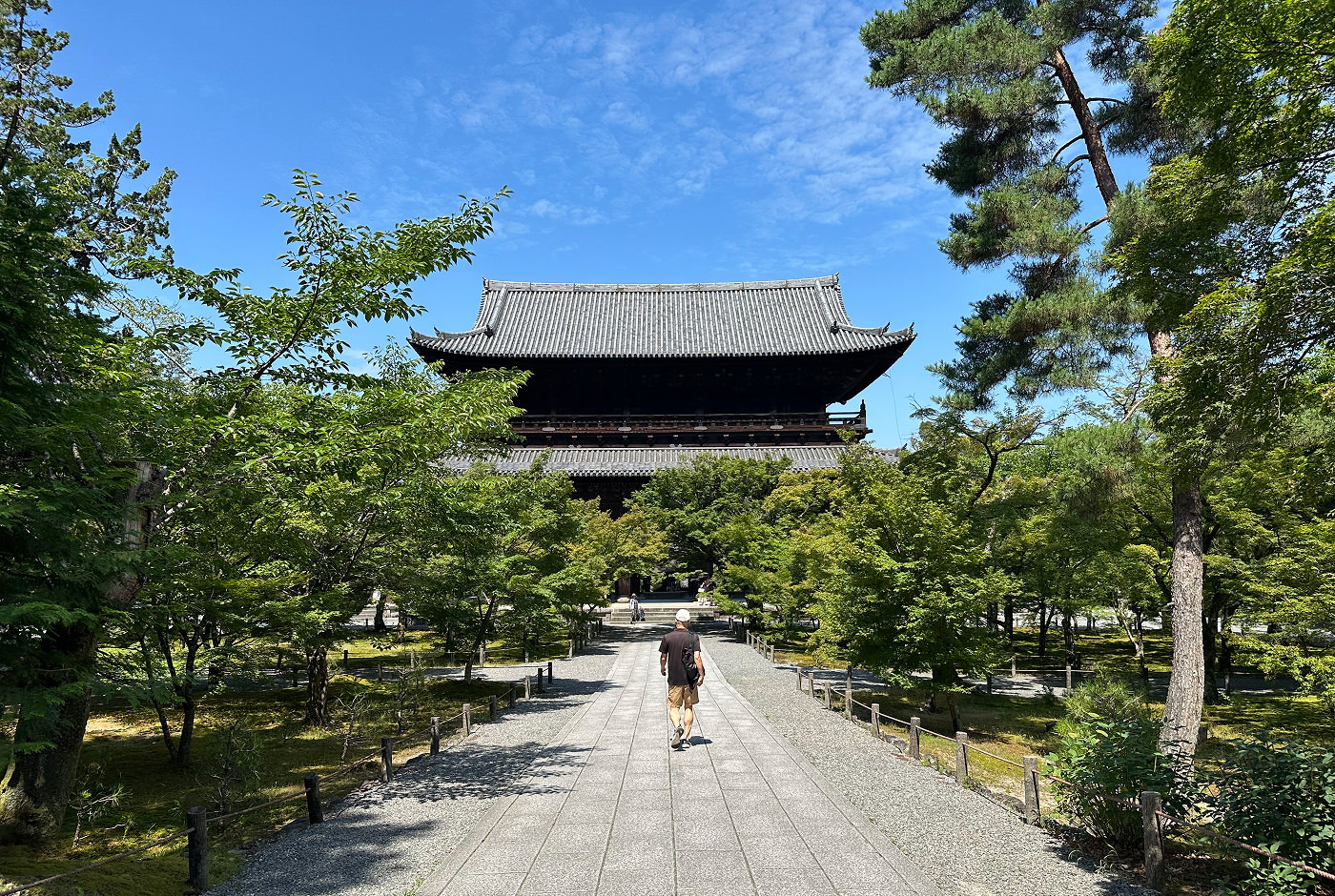 Stort gammalt japanskt tempel med böjt tak och två våningar. En man går på en ljus stengång mot templet. Gången är omgiven av buskiga träd.