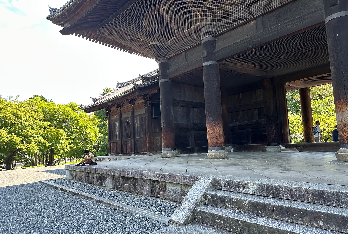 Upphöjt ljust stenfundament där ett stort gammalt japanskt tempel står. En man sitter på fundamentet. Templet är byggt av mörkbrunt trä och har flera pelare.