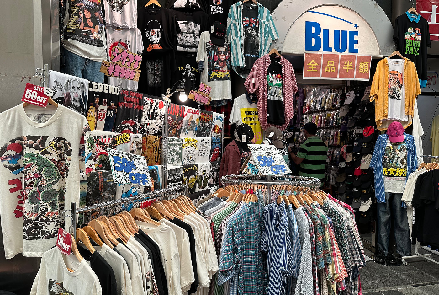 En liten butik fylld till bredden med ljusa och mörka t-shirts ed tryck. En skylt ovanför ingången lyder "Blue".