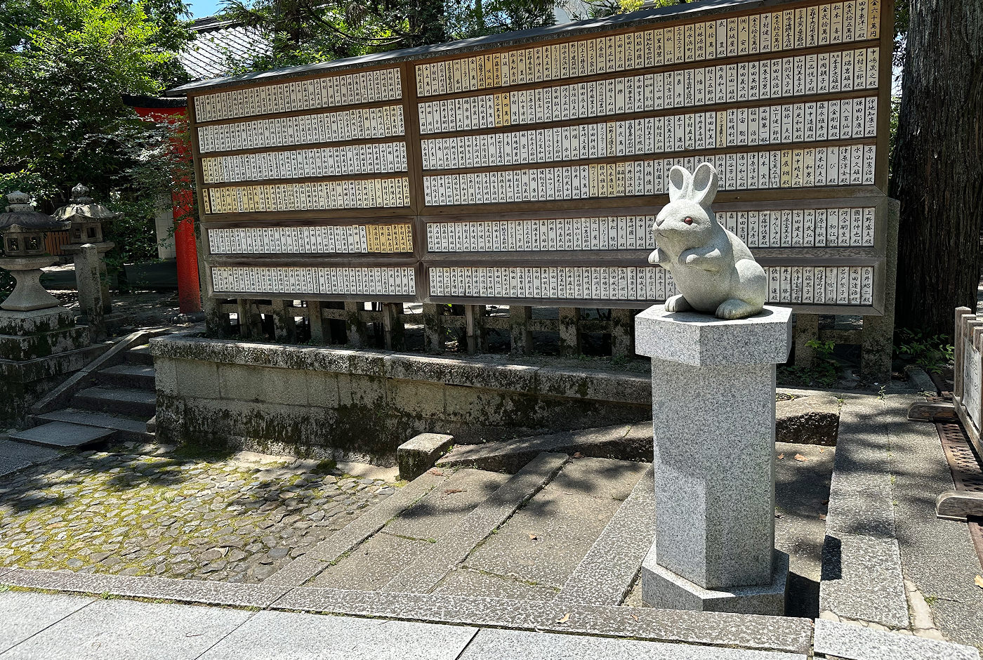 En stenstaty av en kanin på en stenpiedestal. I bakgrunden finns en stor tavla med många små avlånga vita träbitar med japansk skrift på.