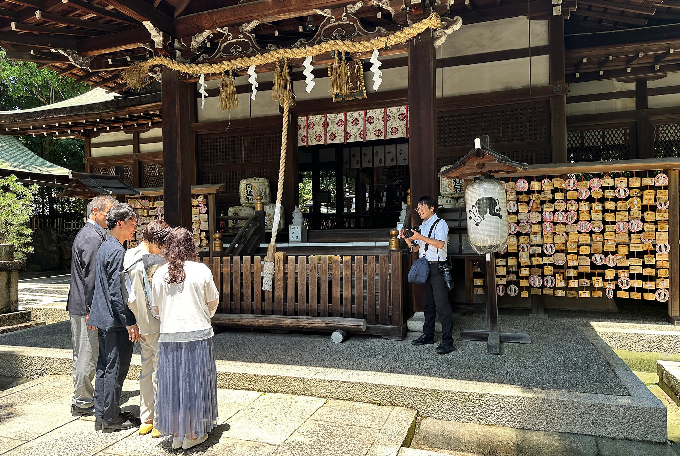 En grupp på fyra människor fotograferas stående vid ett gammalt japanskt trätempel. Fotografen instruerar gruppen.