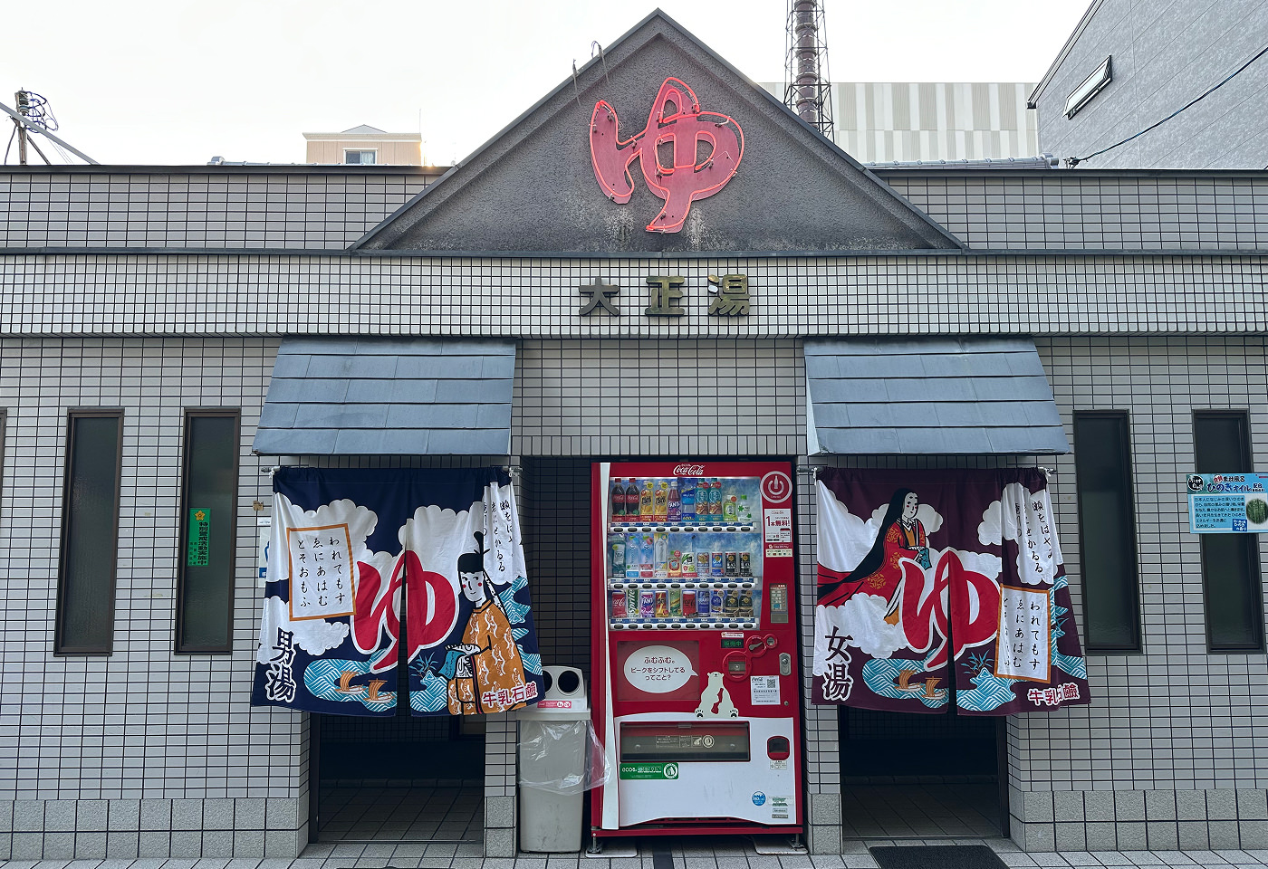 Utanför ett klassikt japanskt badhus. Fasaden består av ljust kakel och har två ingångar, ett med rött och ett med blått skynke. I mitten står en röd dryckesautomat.