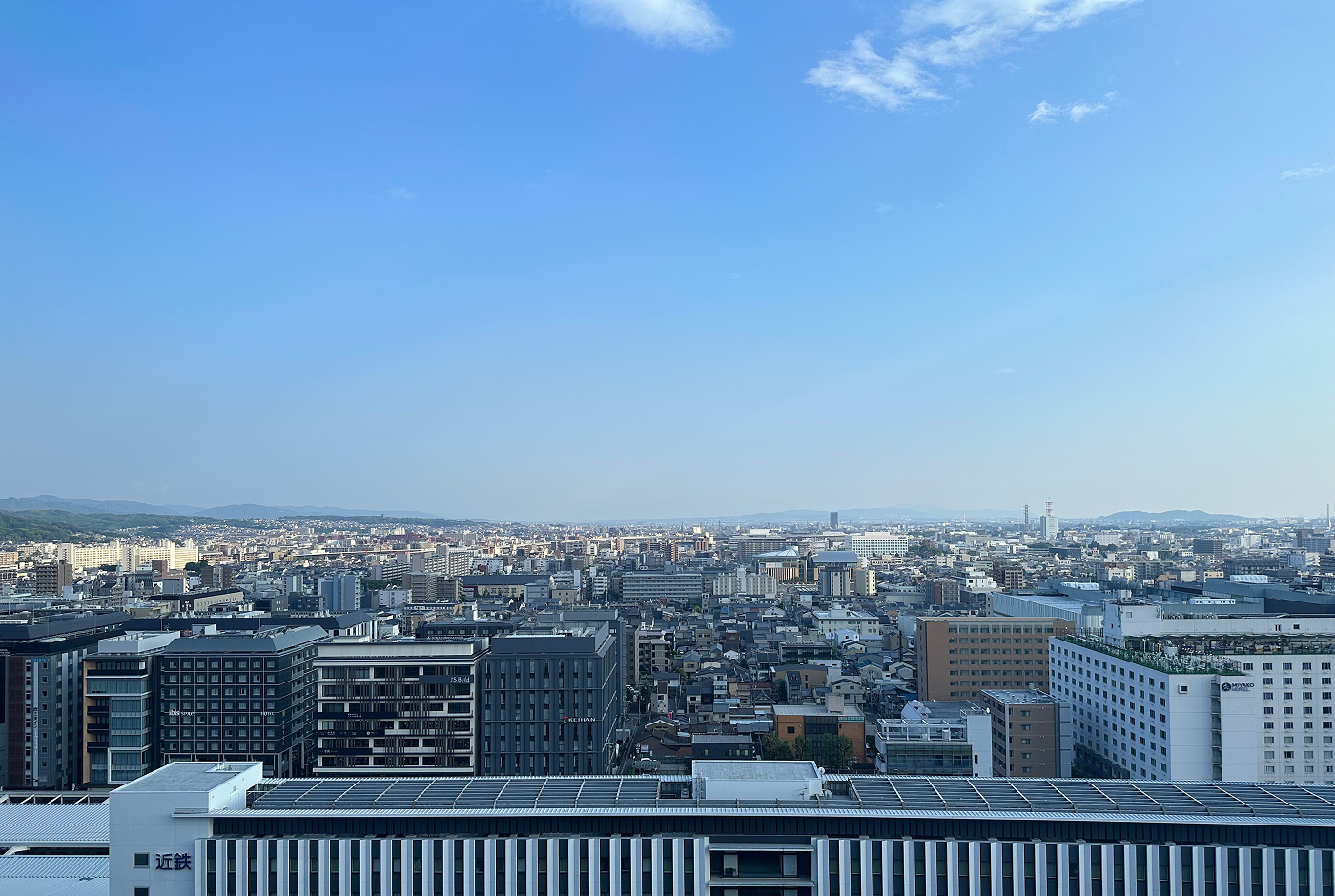 Milsvid utsikt över storstaden Kobe. Långt bort skymtar lummiga berg. Himlen är nästan helt molnfri.