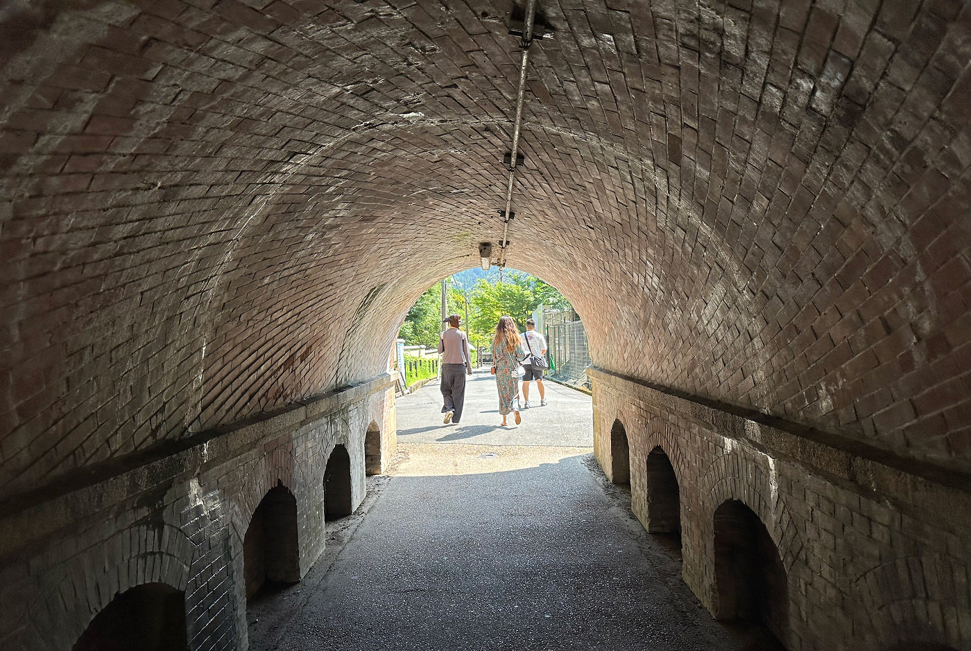 En liten och rund tunnel byggd med brunt tegel. Tre personer har precis gått ut vid den bortre ändan där solen lyser.