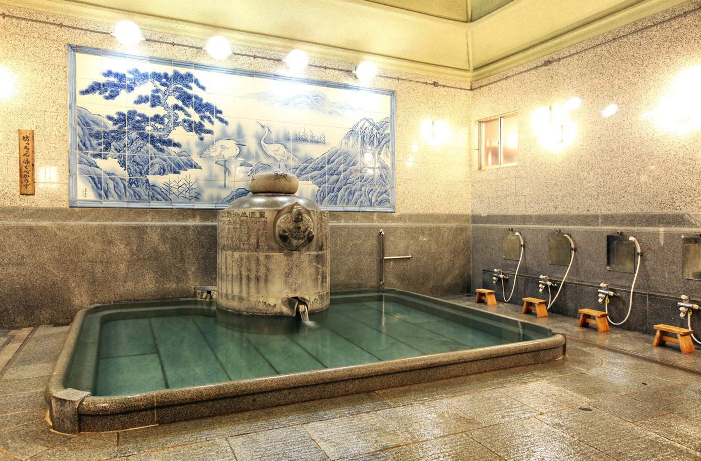 Liten varm pool i ett gammaldags japanskt badhus. Interiören är av sten med en vitblå kakelmålning av fåglar ite i naturen på väggen. Sittduschar med träpallar står till höger.