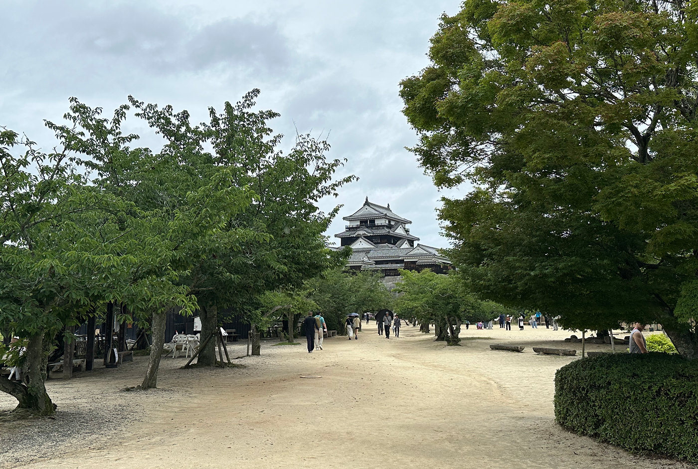 En ljus grusgård med lummiga i träd. I fjärran ser man tornet på Matsuyama Castle.