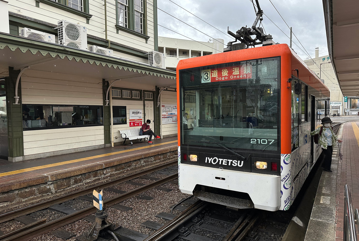 En vit och orange spårvagn vid en station mellan vita hus. Skylten lyder "3 Dogo Onsen".