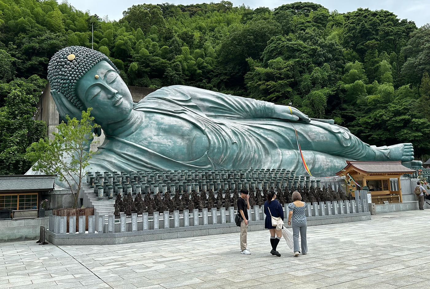 En enorm turkos staty av en liggande buddha på en stor stenyta omgiven av ett lummigt berg. Tre personer står framför statyn.