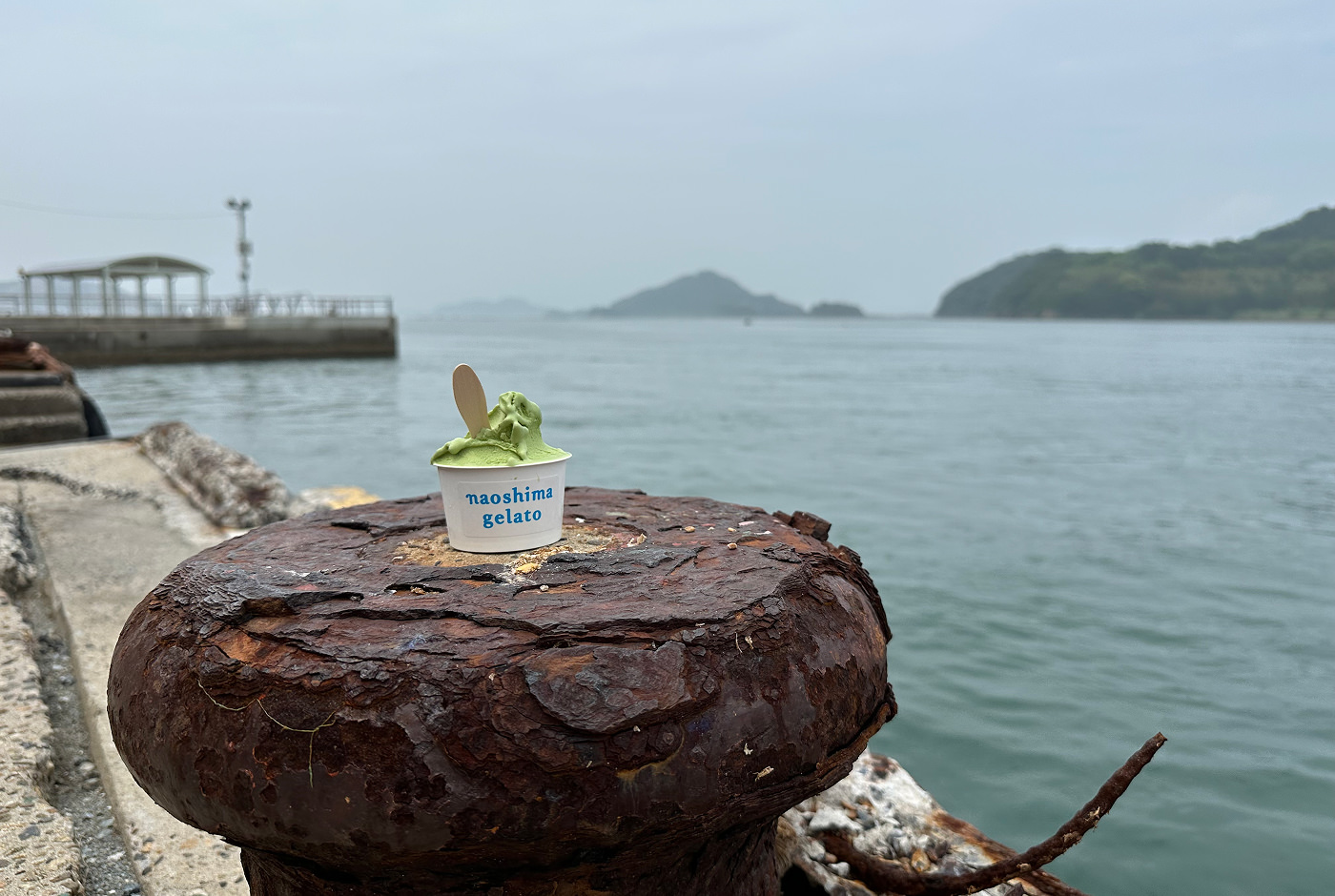 Sliten och rostig pollare vid en hamn. Ovan den stor en bägare med ljusgrön glass med texten "Naoshima gelato".