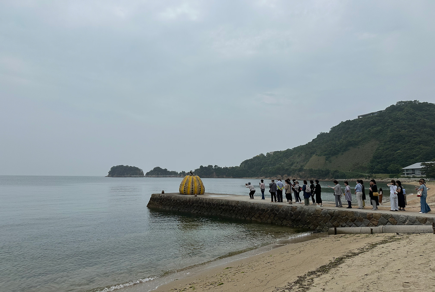 Flera japaner står i kö på en pir vid en strand för att fotografera en stor skulptur av en gul pumpa med svarta prickar.