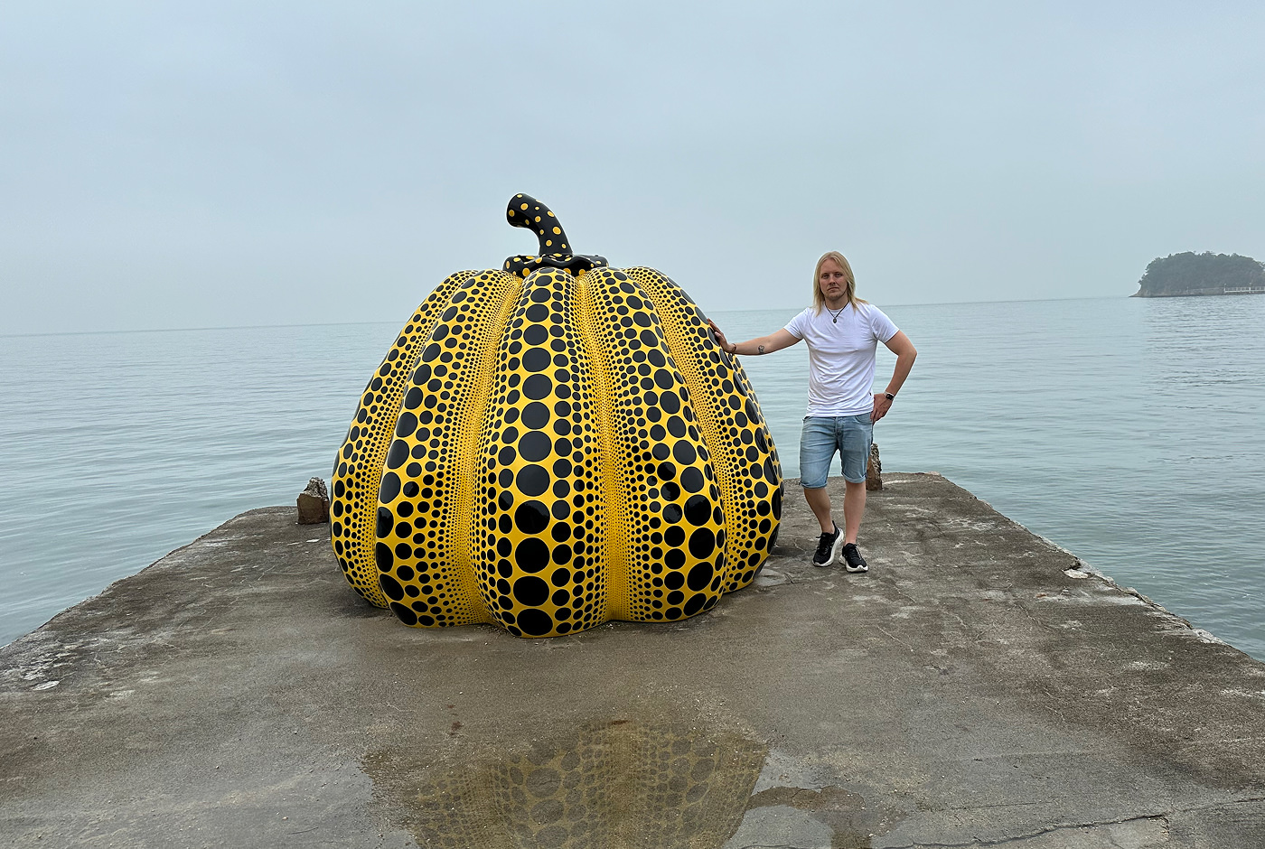 Skulptur av en stor gul pumpa med tusentals svarta prickar. Den står på en betongpir framför ett dimmigt hav. Jag står bredvid den och håller handen mot den.