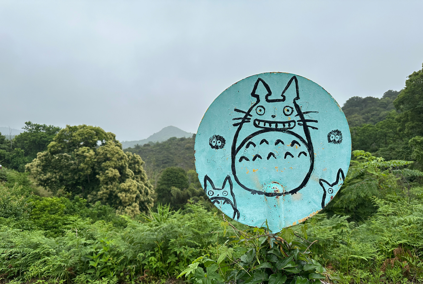 Ljusblå rund skylt med den tecknade figuren Totoro. I bakgrunden lummiga berg.