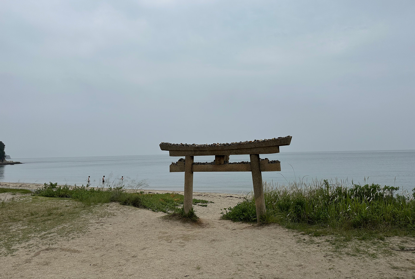 Gråbrun torii-port nere vid en sandstrand. I vattnet badar tre personer.