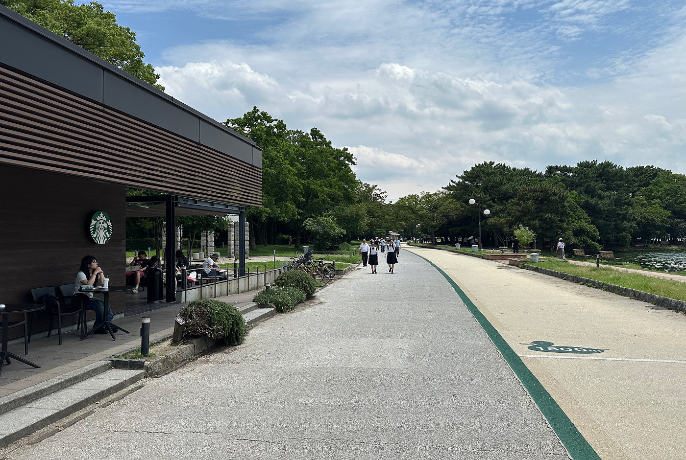 Lång promenadgång i sten genom en lummig park. Till höger skymtas vatten. Till vänster sitter en tjej på en stol vid ett Starbucks-fik.