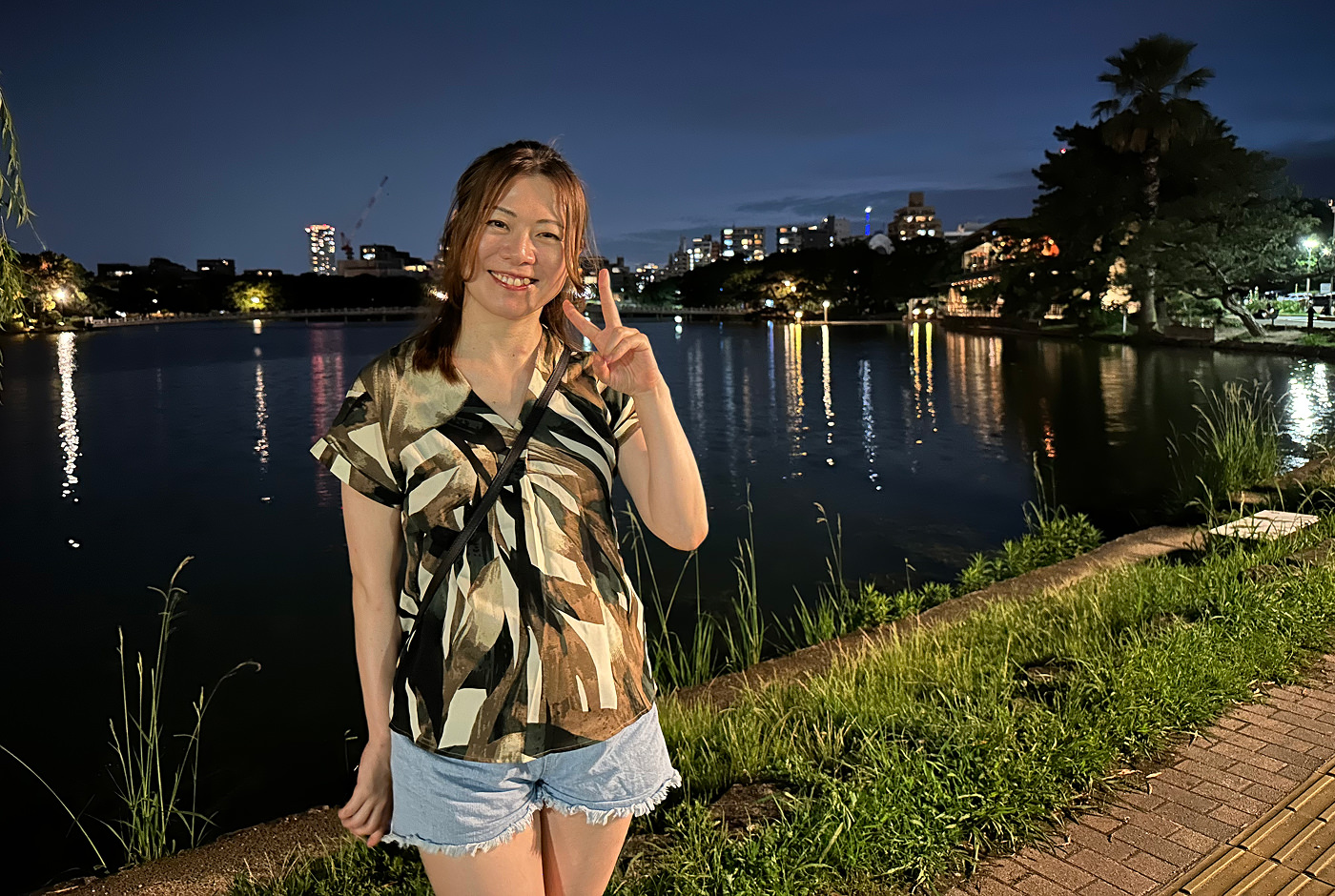 Ung japansk kvinna med shorts och mönstrad skjorta i jordiga färger ler och visar fredstecknet till kameran. bakom henne är en insjö i en stor park. Det är sent på kvällen.