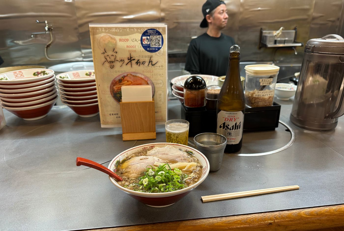 Skål med ramen, ett glas vatten och ett glas öl och dess flaska står på en bardisk omgiven av tallrikar och kryddor.