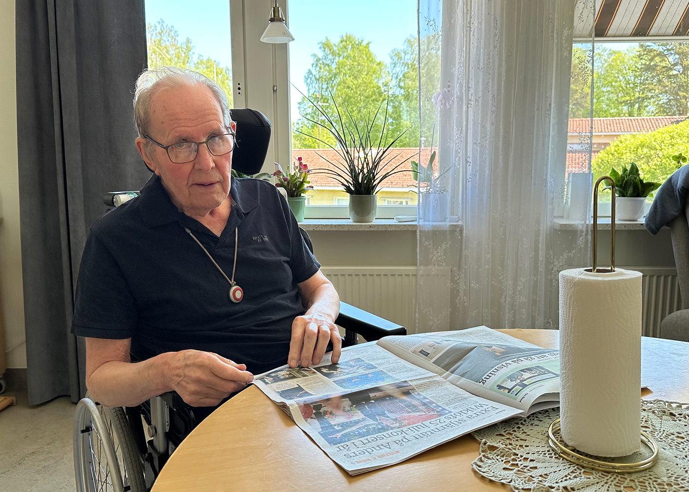 Äldre man med svart kortärmad tröja och glasögon sitter vid ett bord och läser en tidning på bordet.